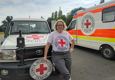 Eine Frau mit einem T-Shirt des Internationalen Komitees vom Roten KReuz steht vor einem IKRK-Fahrzeug. Im Hintergrund ein IKRK-Ambulanzfahrzeug.