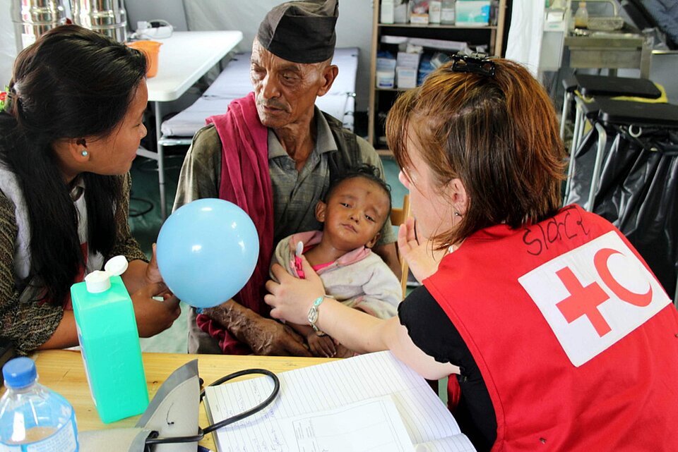 Eine Helferin vom Nepalesischen Roten Kreuz und eine Mitarbeiterin vom Finnischen Roten KReuz behalndeln ein Kind, das von einem älteren Mann gehalten wird.