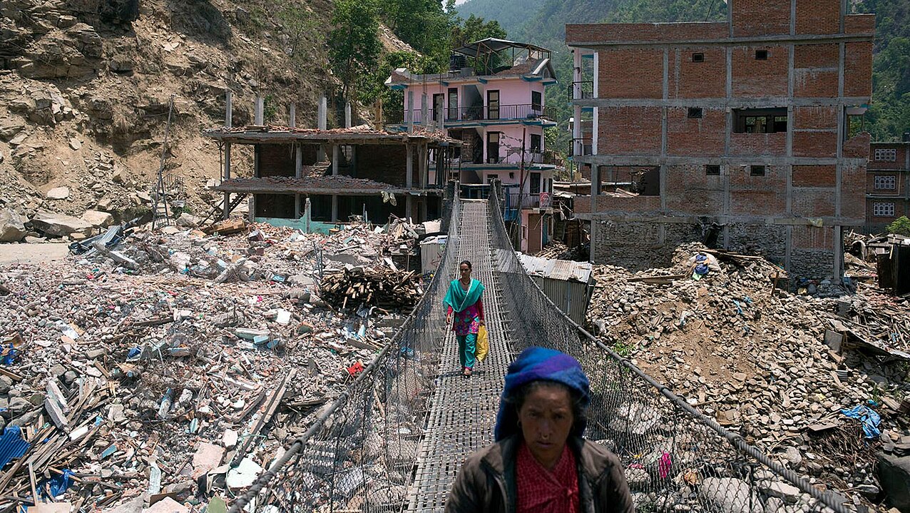 Menschen gehen über eine Hängebrücke, im Hintergrund die zerstörete Ortschaft Singati in Nepal